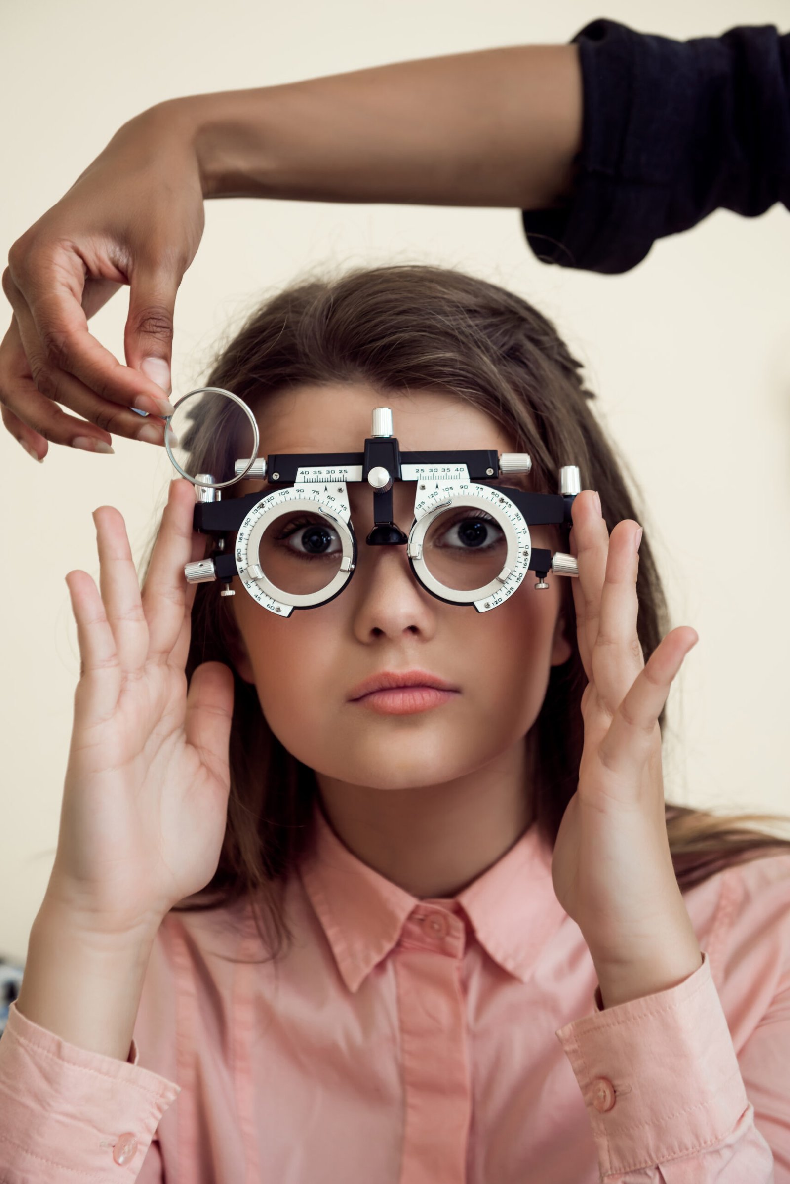 horizontal shot of interested and curious caucasian girl on appointment with eye care specialist wearing phoropter while ophthalmologist checking her vision, sitting over yellow background